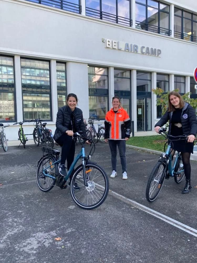 Trois personnes avec des vélos électriques devant le bâtiment Bel Air Camp.