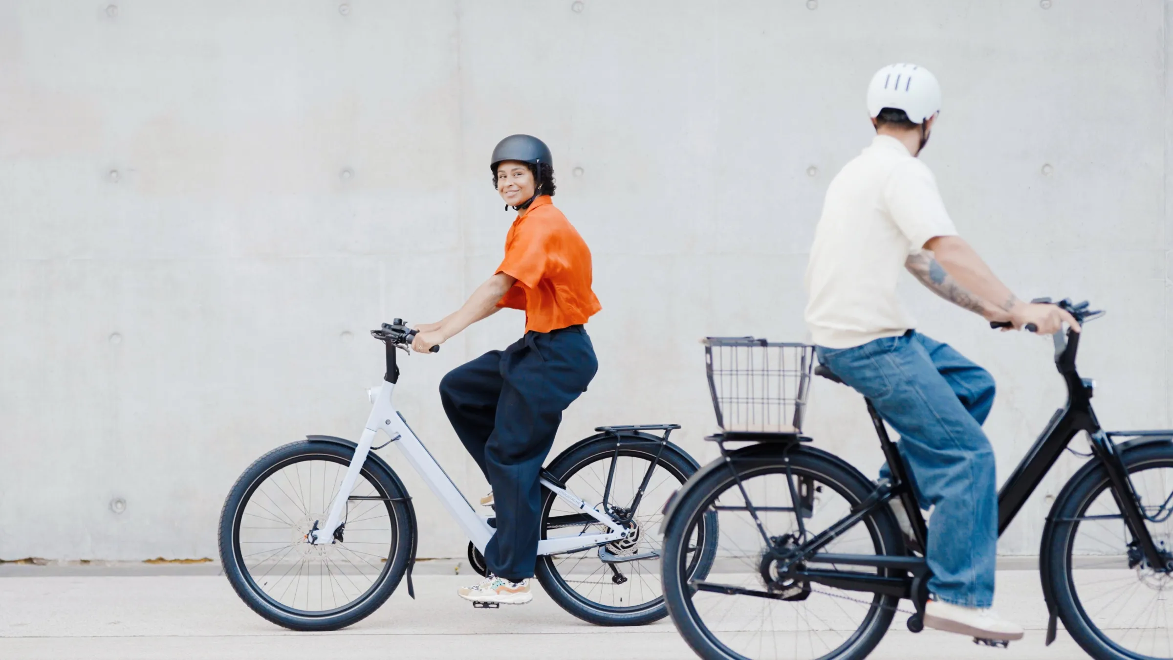 Une femme en chemise orange sur un vélo électrique Moustache blanc se retourne vers un homme casqué qui la suit sur un vélo noir à panier, devant un mur de béton. Photo : Moustache Bikes