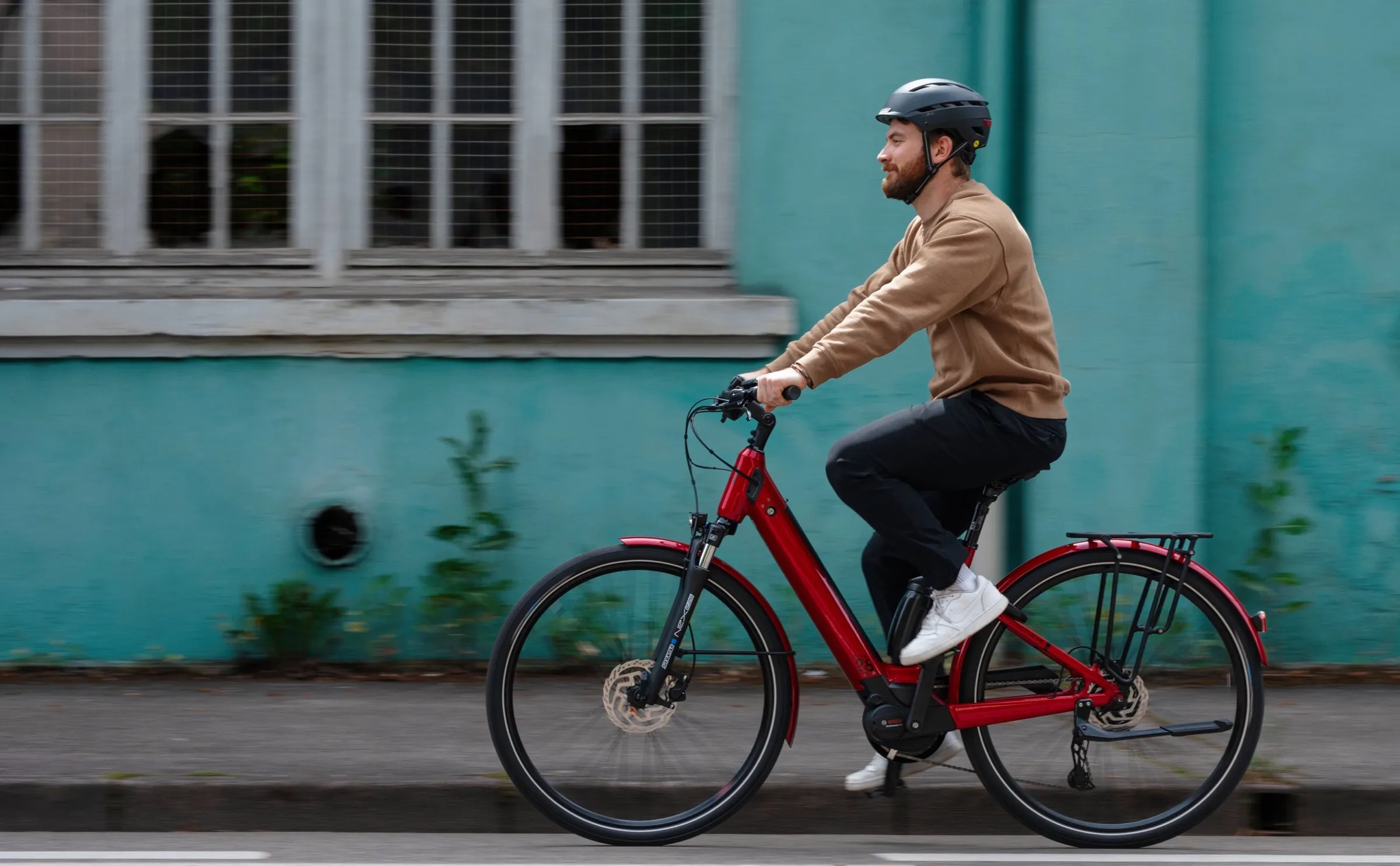 Salarié casqué roulant en ville sur un vélo électrique Moustache Samedi 28 rouge, devant une façade turquoise. Photo : Moustache Bikes