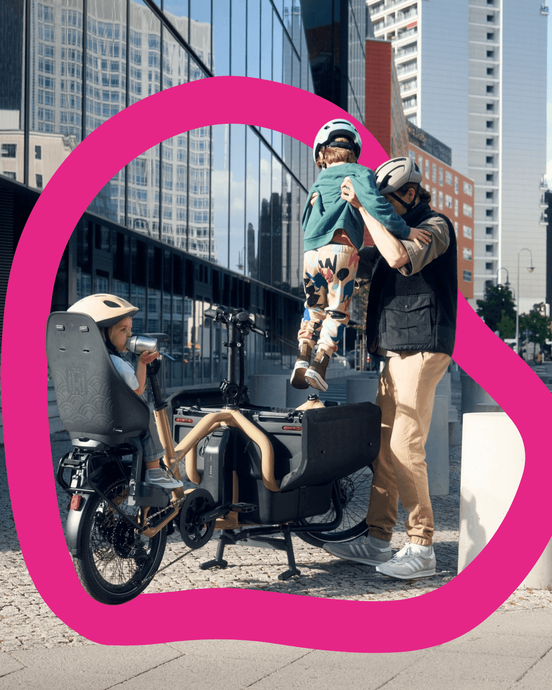 Father lifts his child up while another child sits in a cargo bike, with modern buildings in the background.