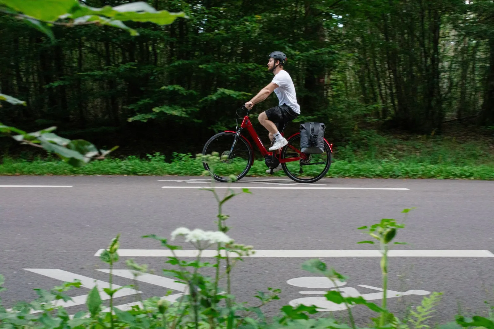 Cycliste casqué sur un Moustache Samedi 28 rouge équipé d’une sacoche, roulant sur une piste cyclable en lisière de forêt.